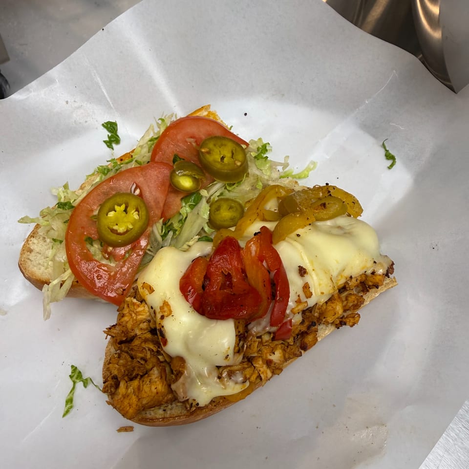 Open-faced chicken sandwich topped with melted cheese, roasted peppers, lettuce, tomatoes, and jalapeños on deli paper.