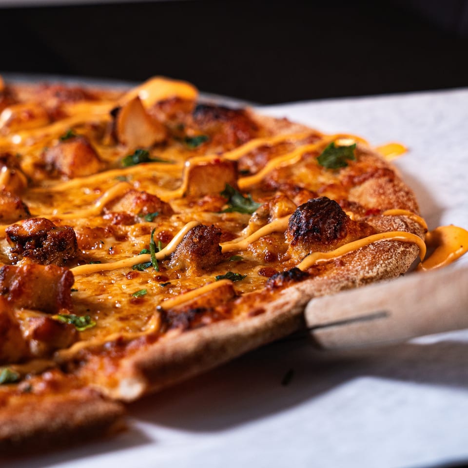 Close-up of a rectangular pizza topped with roasted meat, melted cheese, herbs, and orange sauce drizzle on parchment paper.